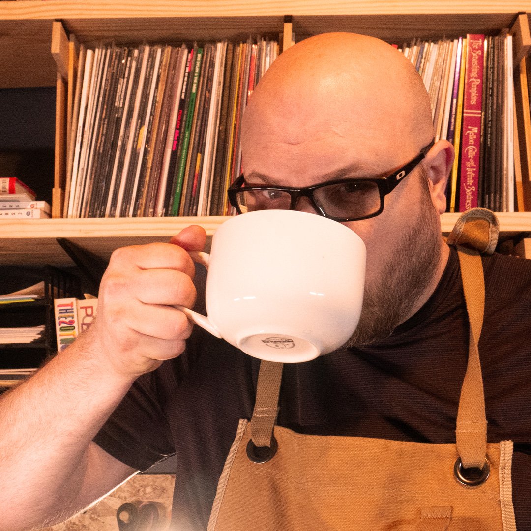 John sipping from an oversized white cup in front of a shelf of records.