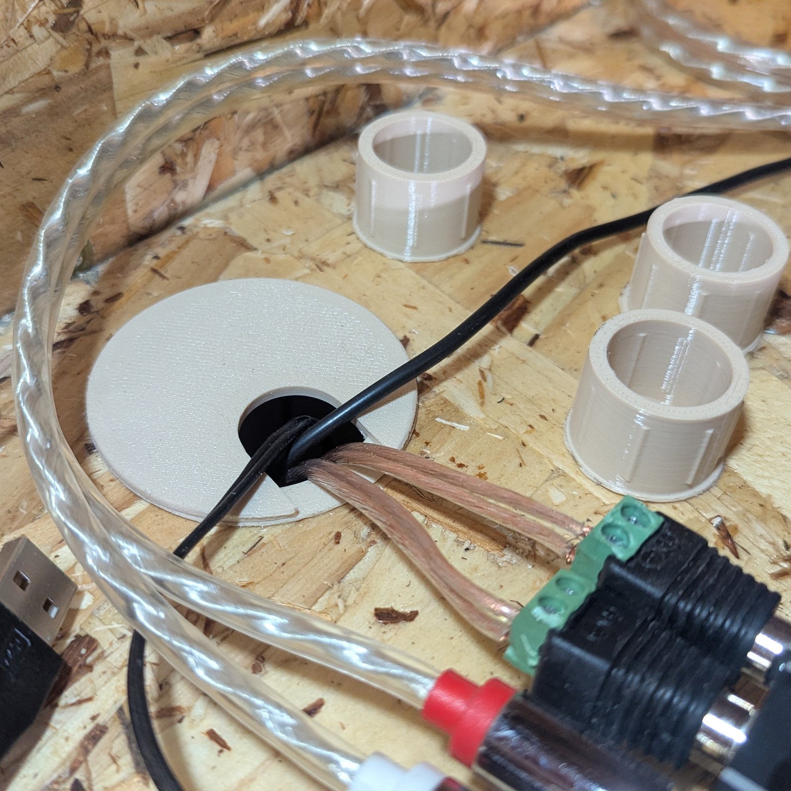 Printed tan desk grommet installed in an OSB desktop with audio and power cables running through it, beside three printed bushings.