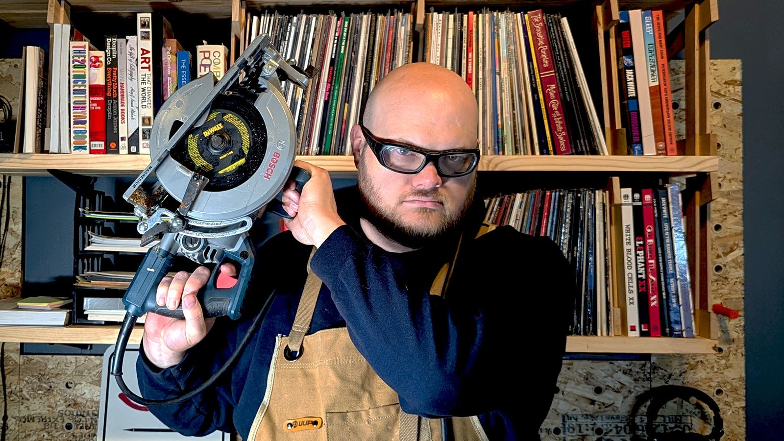 John in safety glasses and a canvas apron, hoisting a circular saw onto his shoulder in front of shelves of records and books.