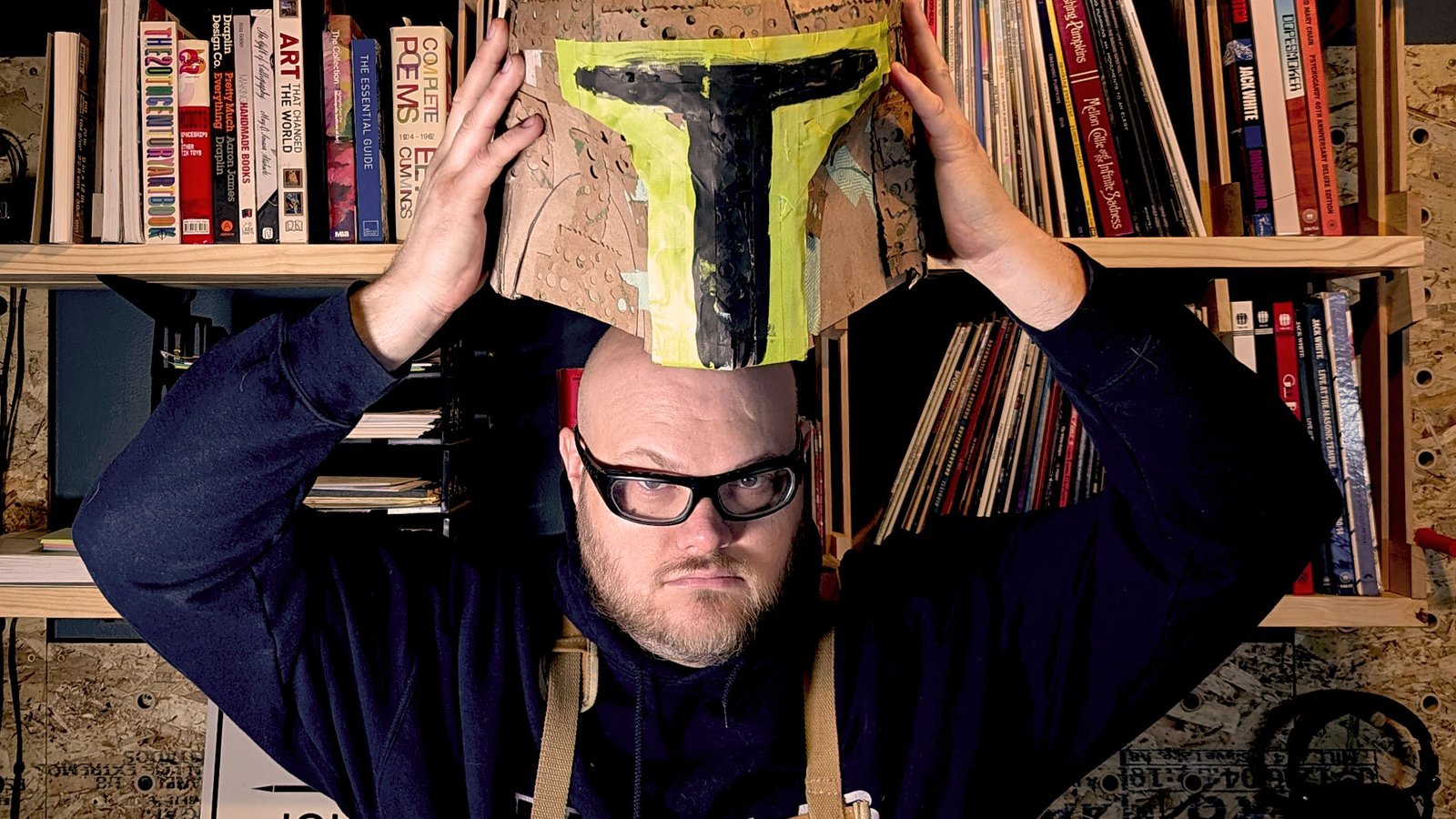 John holding a handmade cardboard bounty-hunter helmet, painted yellow and black, above his head.